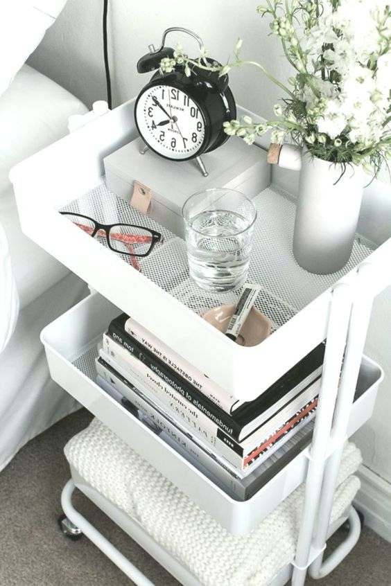 White 3-tier rolling cart used as a nightstand with books, a clock, and a vase of flowers beside a bed in a small apartment bedroom