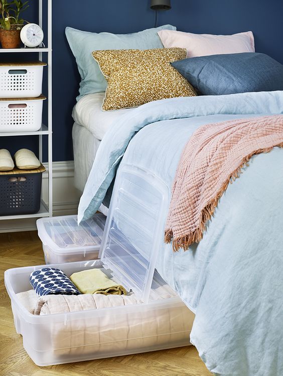 Clear plastic under-bed storage bins being pulled out from beneath a bed with blue and neutral bedding in a small apartment bedroom