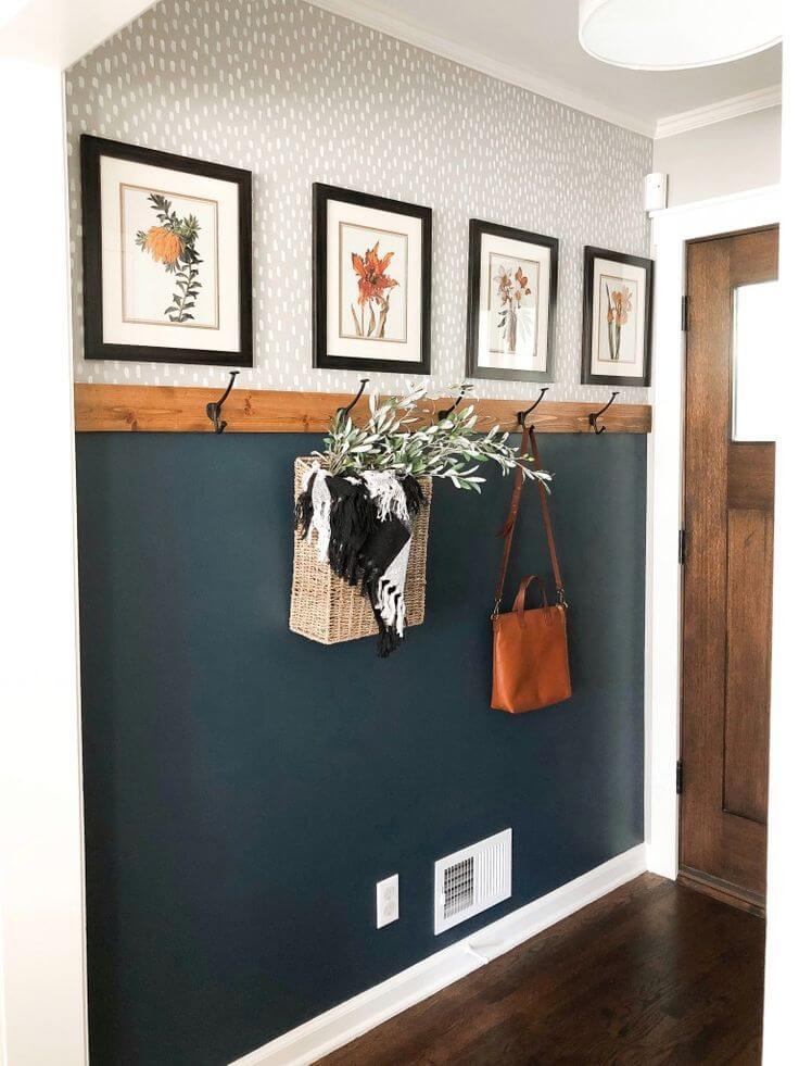Small apartment entryway with a two tone accent wall, patterned wallpaper on top, dark teal on the bottom with a wood hook rail, botanical frames and hanging bags