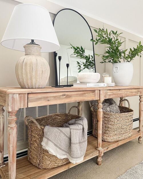 Small apartment entryway with a textured stone table lamp, arch mirror, white vase with green stems and woven baskets underneath on a rustic wood console table
