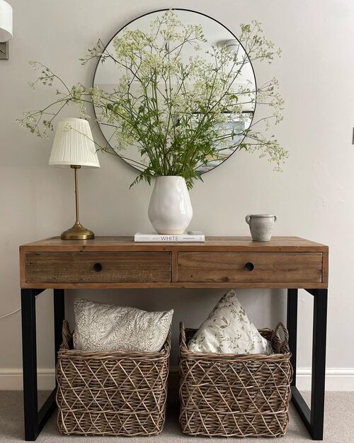 Small apartment entryway with two large woven baskets under a wood console table, round black mirror, white vase with wildflowers and a brass lamp