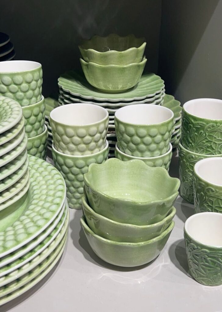 Stacked sage green textured ceramic plates, bowls, and mugs on a shelf