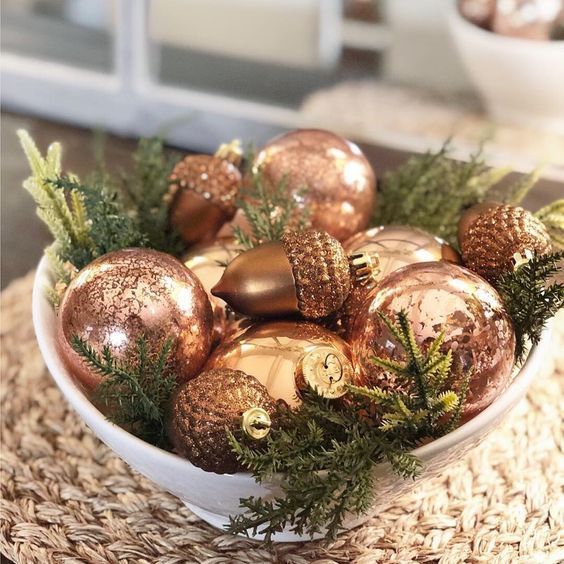 Copper and rose gold ornaments filling a white bowl with greenery for a winter centerpiece