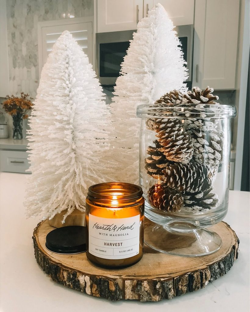 Frosted white mini trees beside a glass jar of pinecones and a lit candle winter centerpiece