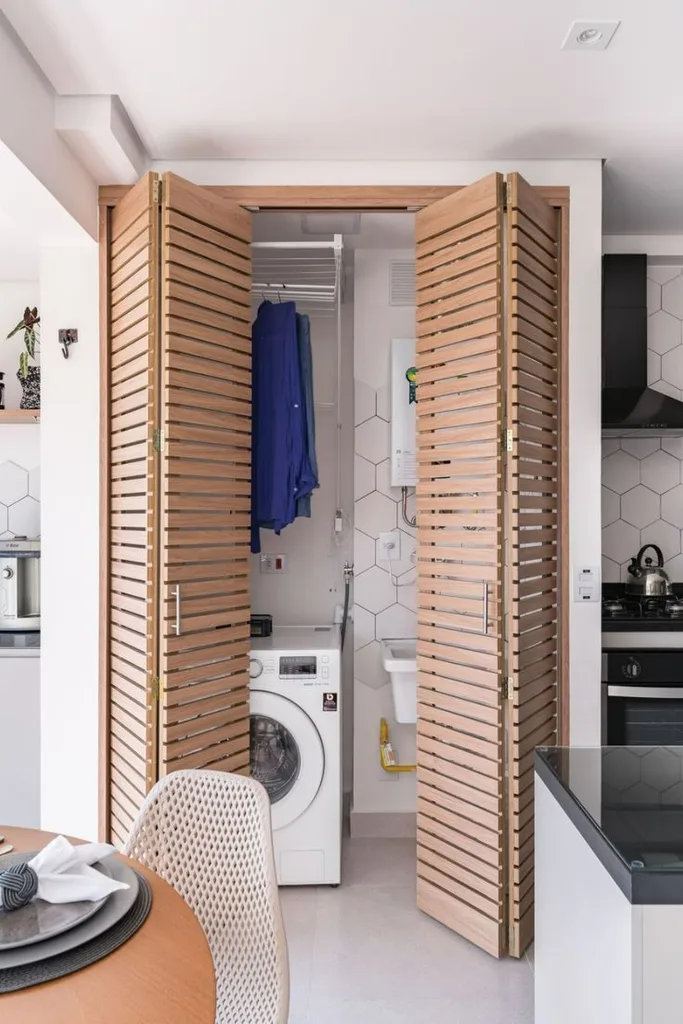 Slatted wood bifold doors open on a small laundry closet next to a kitchen, showing a white washer, blue shirts on a drying rod and hexagon wall tile.
