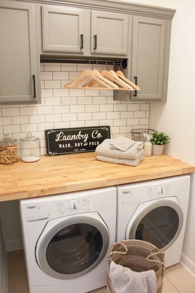 Butcher block countertop over a side-by-side washer and dryer, with folded towels, glass jars and a laundry sign, and gray cabinets above.