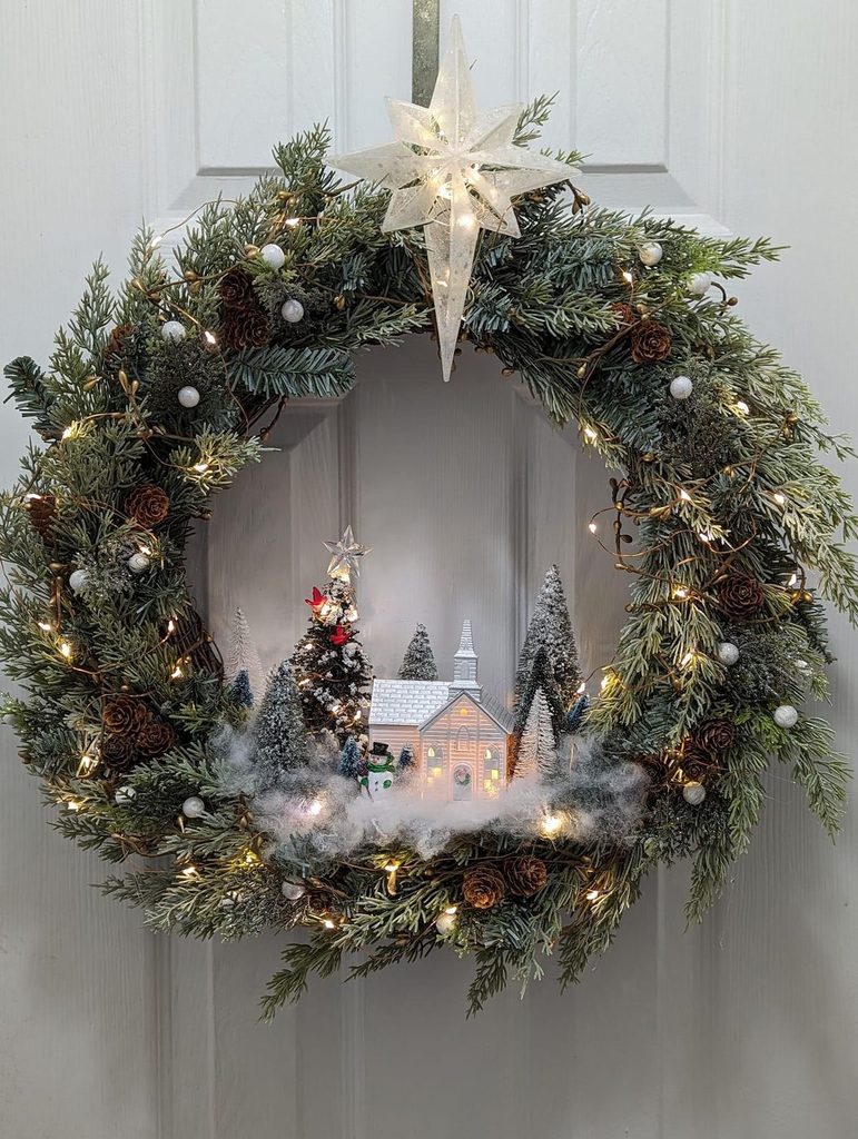 Evergreen village wreath with one lit white house and bottle brush trees inside the ring, hanging on a door