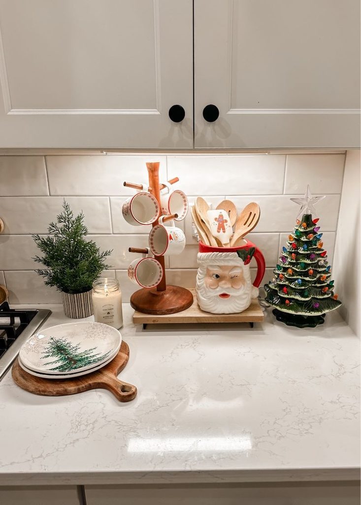 Santa face mug holding wooden spoons beside a mug tree and a retro ceramic tree on a kitchen counter