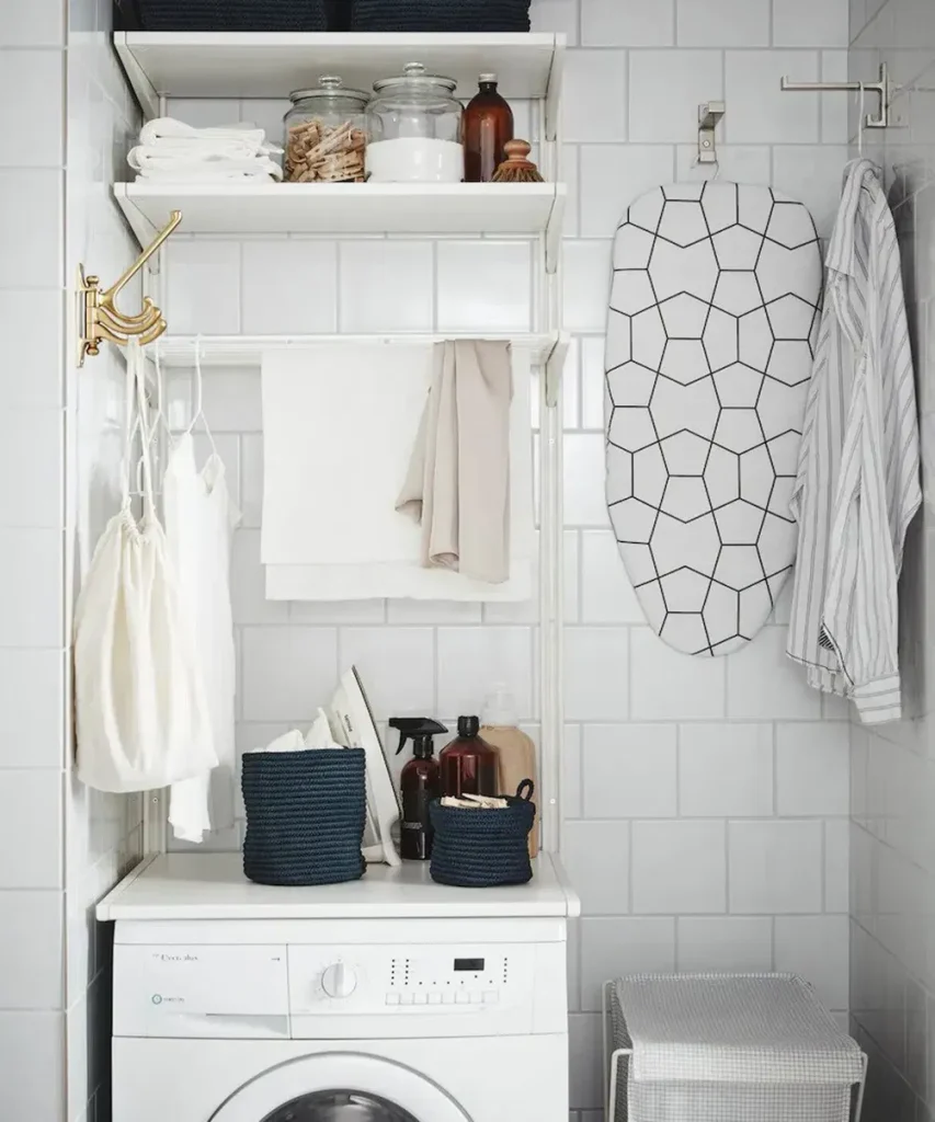 White wall-mounted rail shelves above a washer holding glass jars and towels, with a drying bar, hooks and a hung ironing board on white tile walls.