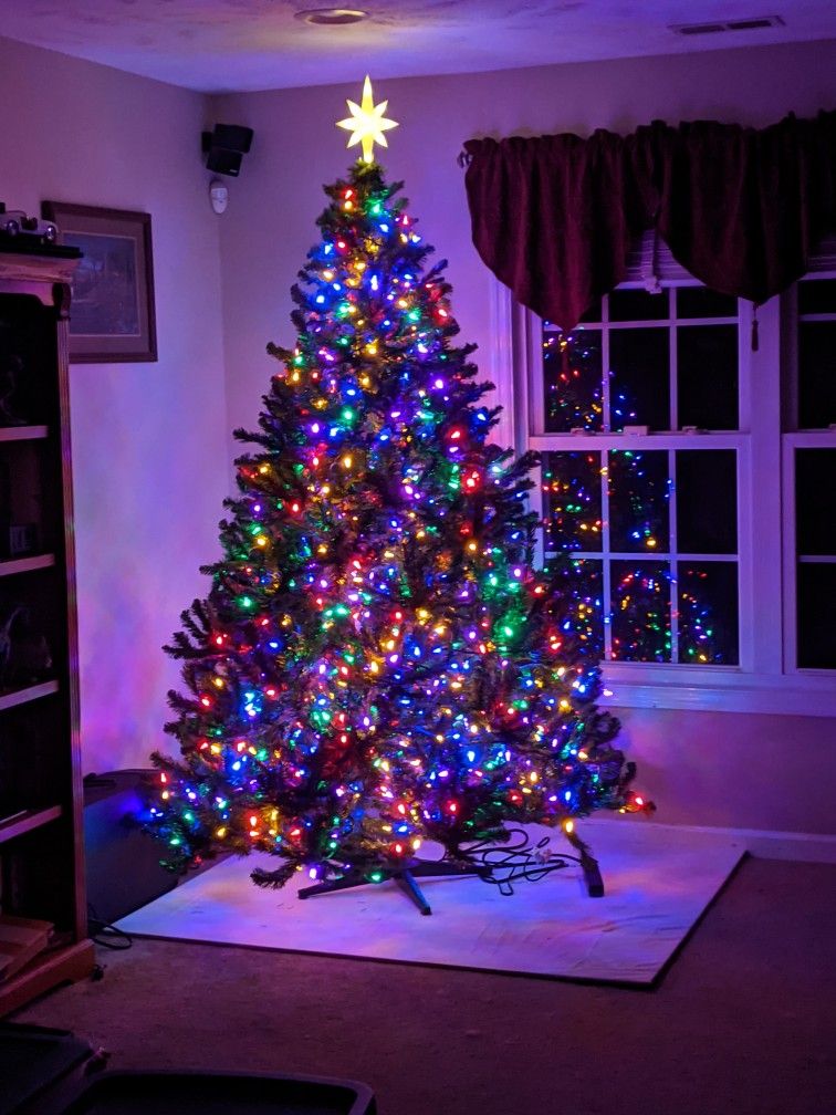 Green Christmas tree with multicolor lights and a star topper in a living room corner.