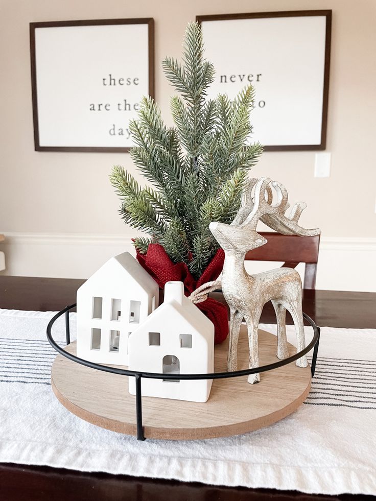 White ceramic houses, a silver reindeer and a mini pine tree on a round wood tray with a black rim