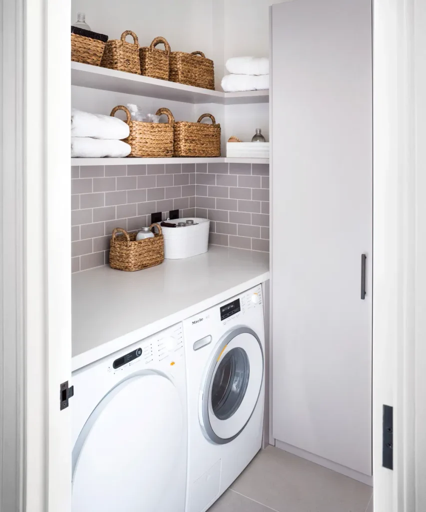 Small apartment laundry closet with a white side-by-side washer and dryer under a counter, wicker baskets on open shelves and gray subway tile behind.