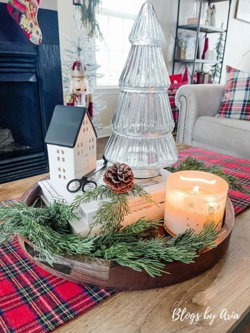 Glass Christmas tree, white house and candle on a book stack in a wood tray on a plaid runner