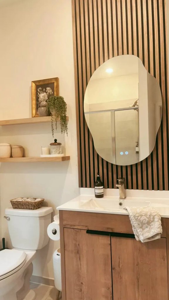 Vertical wood slat accent wall behind an oval mirror in a small bathroom with floating shelves and a toilet